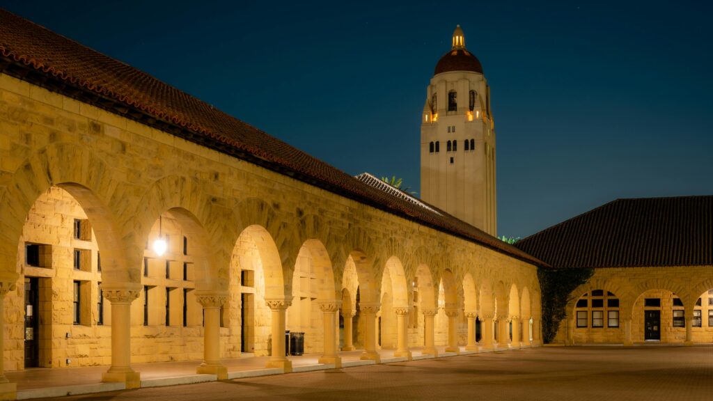 Illuminated Hoover Tower and arches at Stanford University under a dark sky.